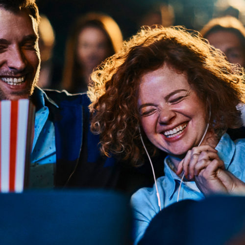 Een lachende man en vrouw in de bioscoop met een bak popcorn. De vrouw heeft oortjes in.