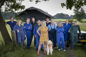 Groepsfoto van alle boeren die dit seizoen meedoen aan Boer zoekt vrouw. De boeren dragen blauwe overalls. In het midden staat Yvon Jaspers in een gele jurk met haar hond.