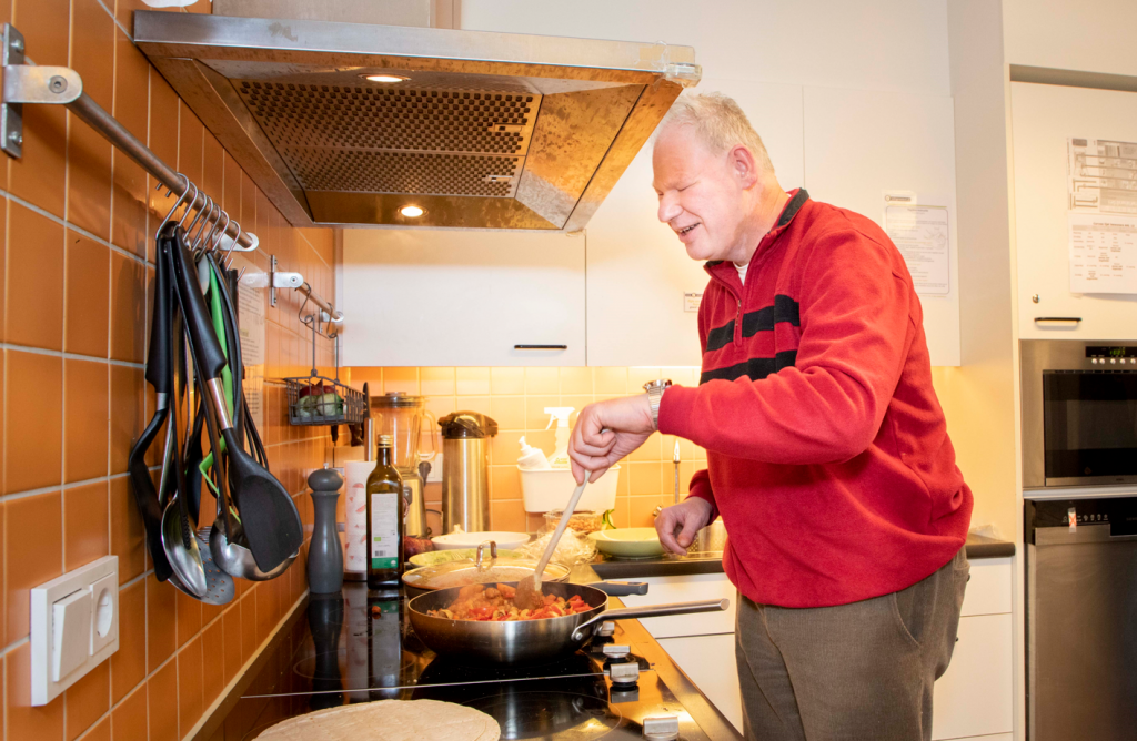 Een bewoner van Bartiméus roert staat in een keuken in een pan te roeren.