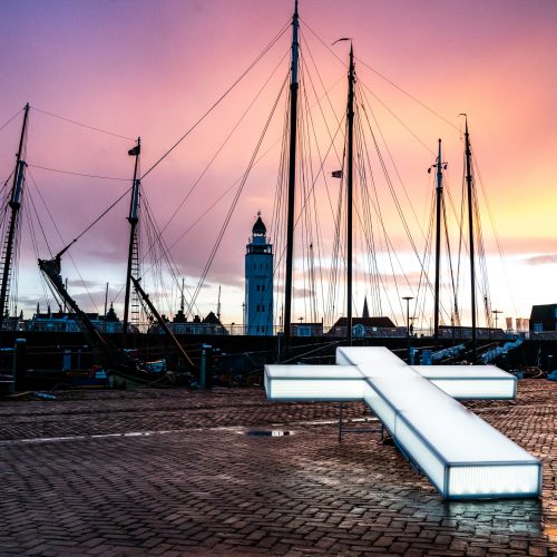 Het grote verlichte kruis van The Passion ligt op de kade van Harlingen Haven. Op de achtergrond masten van boten tegen een oranje-paarse lucht.