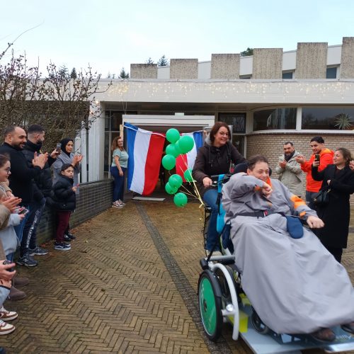 Begeleidster fietst met een bewoner in een rolstoel op een rolstoelfiets door een erehaag van mensen.