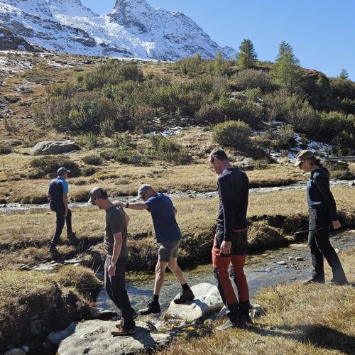 Simon steekt samen met een buddy een beekje over in de Alpen. Er zijn nog 3 wandelaars.